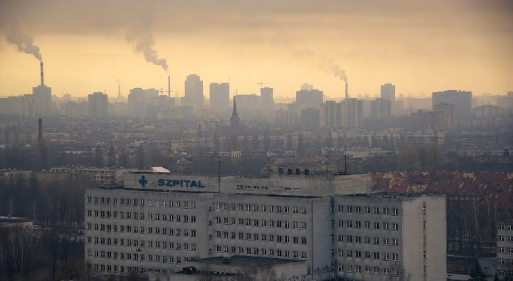 Fotografia wielkiego betonowego szpitala z niebieskim napisem SZPITAL na tle zamglonej, smogowej panoramy miasta z dymiącymi kominami